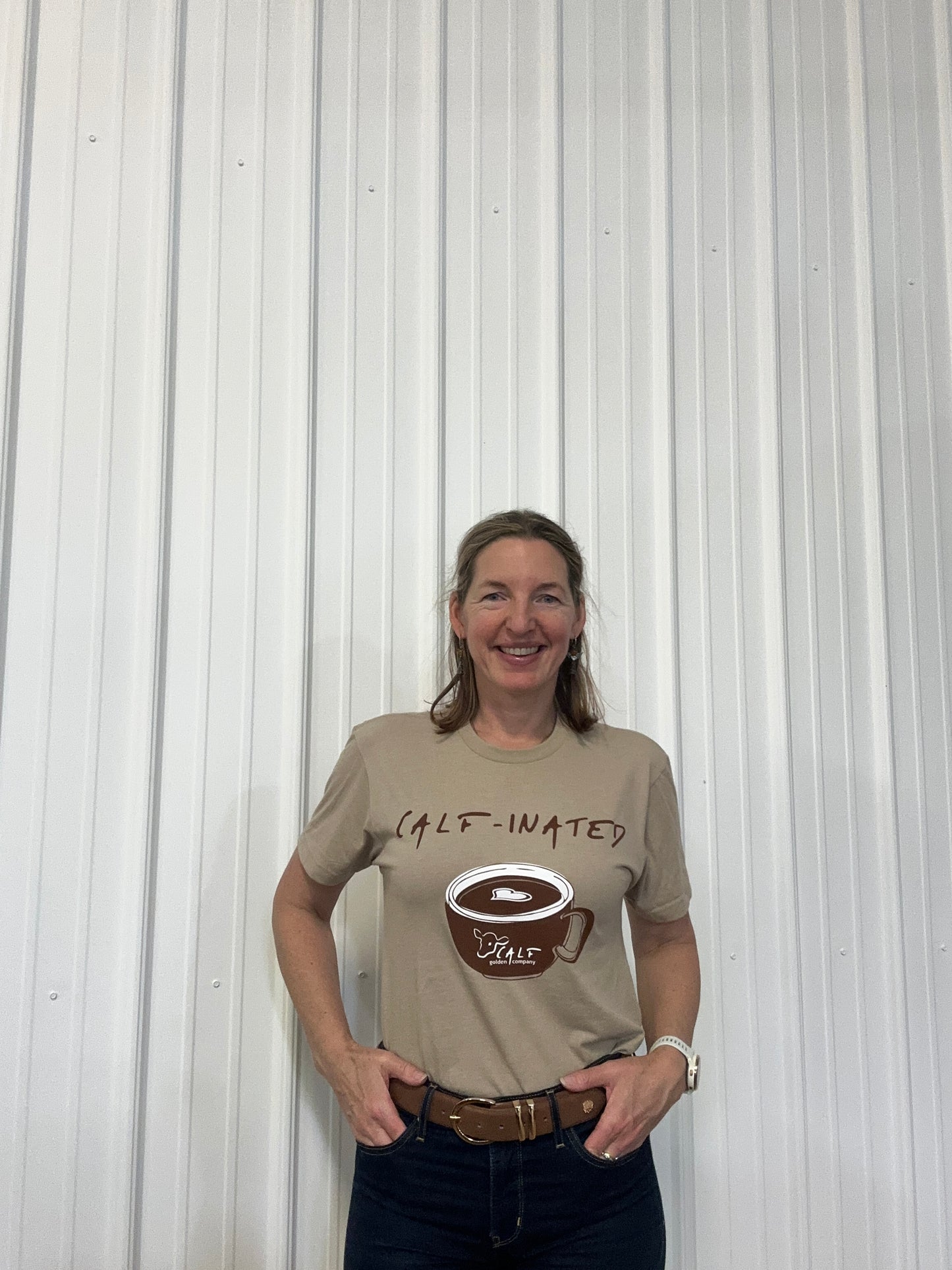 Person wearing a beige t-shirt with a logo and text Calf Hero shirt funny calf-inated with a cup of coffee with an image of a calf on on the coffee mug., standing against a white corrugated background